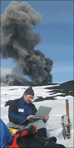 Volcanologist Marco Neri during the winter of 2008-2009 downloads data onto a laptop from the ERN1 radon sensor at the site (later buried in lava) known as the Tower of the Philosopher. Behind him, less than 1 kilometer away, ash billows fro