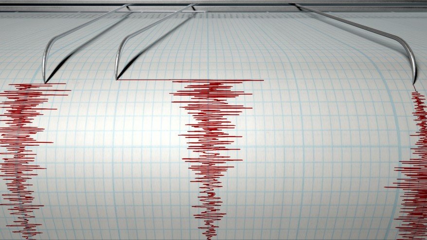 A closeup of a seismograph machine needle drawing a red line on graph paper depicting seismic and eartquake activity 