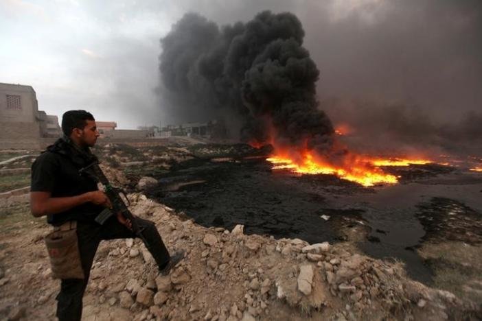 Click to enlarge Iraqi security forces stands with his weapon as fire and smoke rise from oil wells