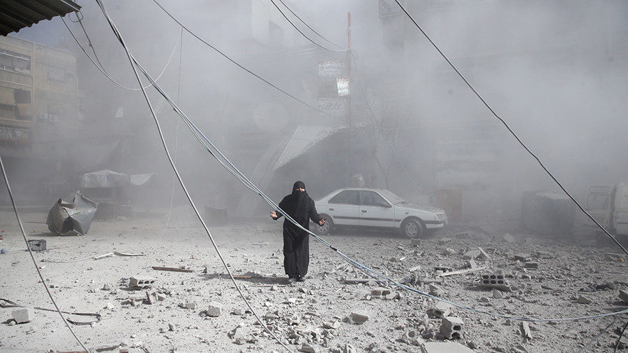 A woman gestures as she walks on rubble of damaged buildings