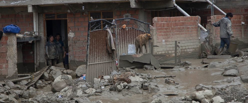 Rocks and mud clog a street outside a flooded home, where a couple and a dog stand by, after a river overflowed in Tiquipaya near Cochabamba, Bolivia, Wednesday, Feb. 7