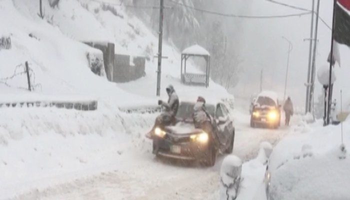 Click to enlarge Two people sit on their car amid thick layers of snow.