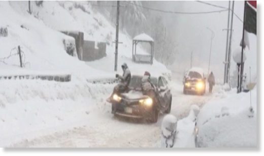 Two people sit on their car amid thick layers of snow.