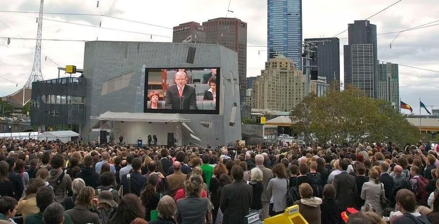 Click to enlarge People gathered listening to a politician on a screen